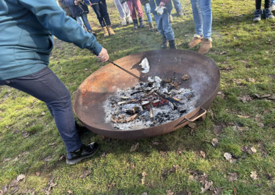 Feuerangebot 2026 - In einer Feuerschale ist Asche und die letzten Reste Glut. Eine Frau geht mit einem Schürhaken durch die Glut.