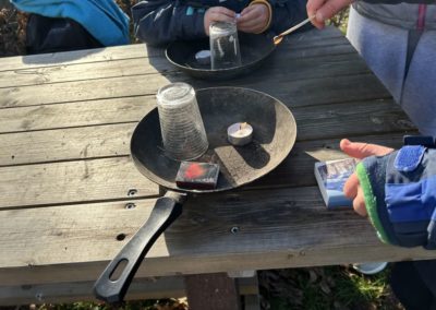 Feuerangebot 2026 - Auf einem Tisch steht eine alte Pfanne. Darin Streichhölzer, ein Teelicht und ein Glas. Es wird gezeigt wie die Flamme des Teelichtes ohne Sauerstoff erlischt.